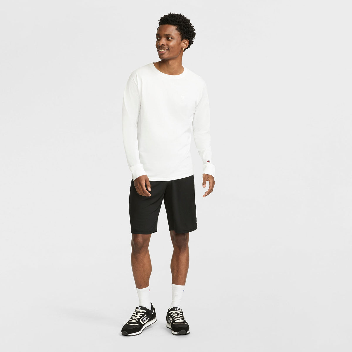 A man smiles, wearing a Champion Classic Long Sleeve T-Shirt with an embroidered C logo, black shorts, white socks, and black sneakers against a plain white background.