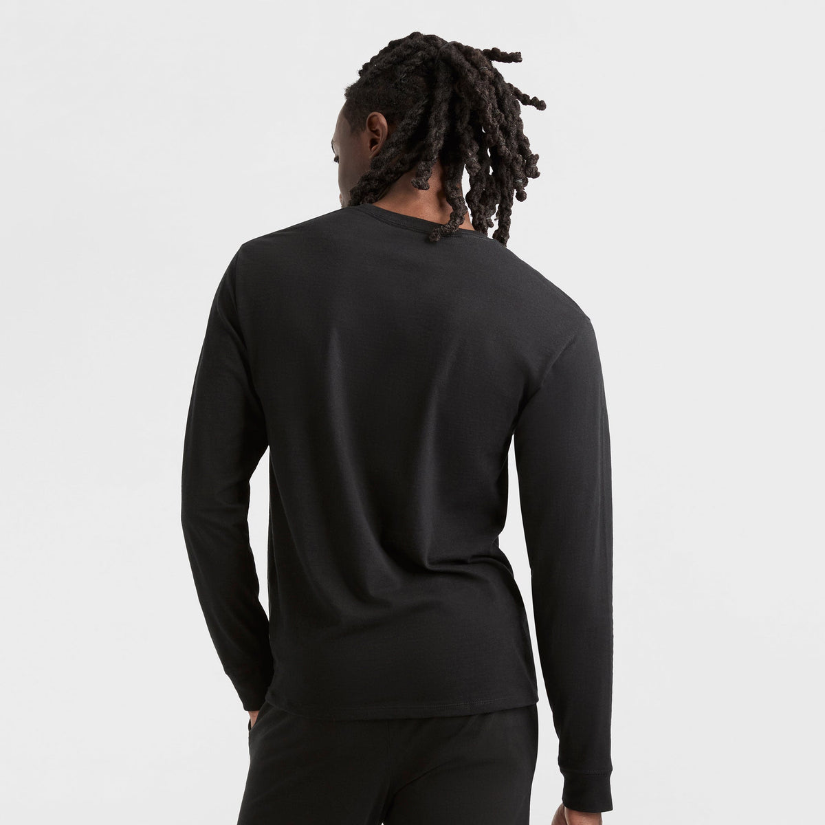 A person with long, twisted hair stands facing away from the camera in a black Champion Classic Long Sleeve T-Shirt with Embroidered C Logo and black pants, set against a plain white background.