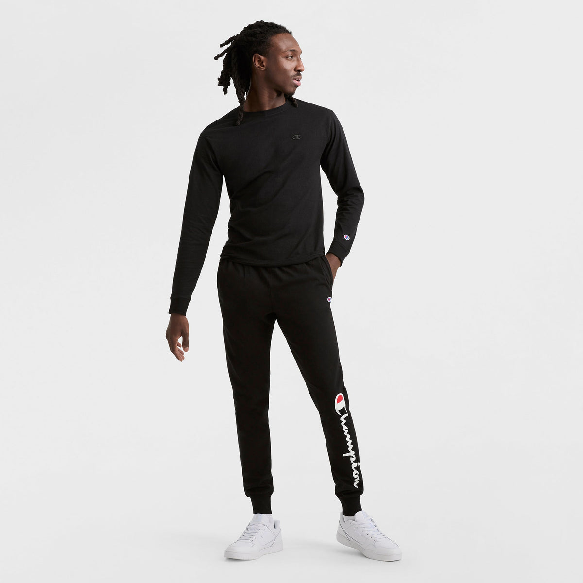 A man with long braided hair wears a black Champion Classic Long Sleeve T-Shirt with an embroidered C logo and black joggers, paired with white sneakers, standing against a plain white background.