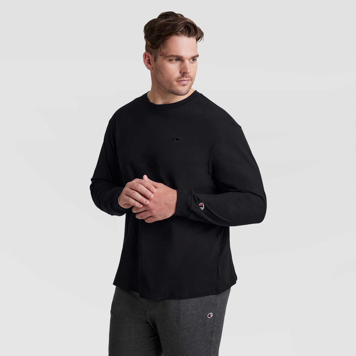 A man wearing the Champion Classic Long Sleeve T-Shirt with Embroidered C Logo (Big & Tall) in black and dark gray pants stands against a plain light gray background, looking to his left with his hands lightly clasped.