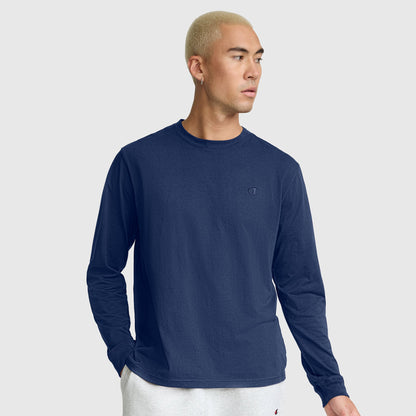 A person with short blond hair wears a navy blue Champion Classic Long Sleeve T-Shirt with an embroidered C logo, made from a soft cotton blend, along with light gray sweatpants. They stand against a plain light gray background, one hand in their pocket and looking to the side.