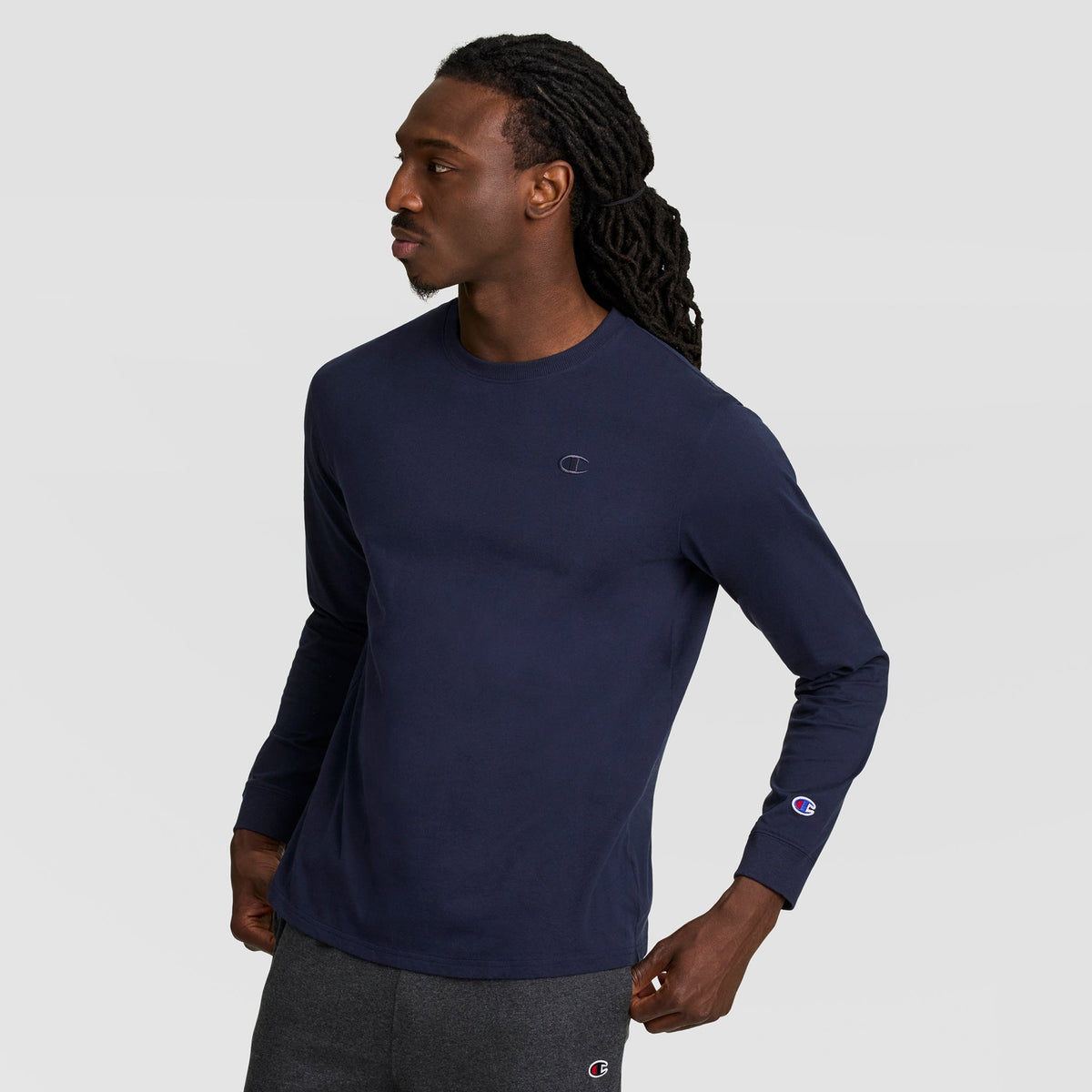A man with long braided hair wears a navy Classic Long Sleeve T-Shirt with an embroidered C logo from Champion and dark gray pants, looking to his left with hands on hips against a plain light gray background.