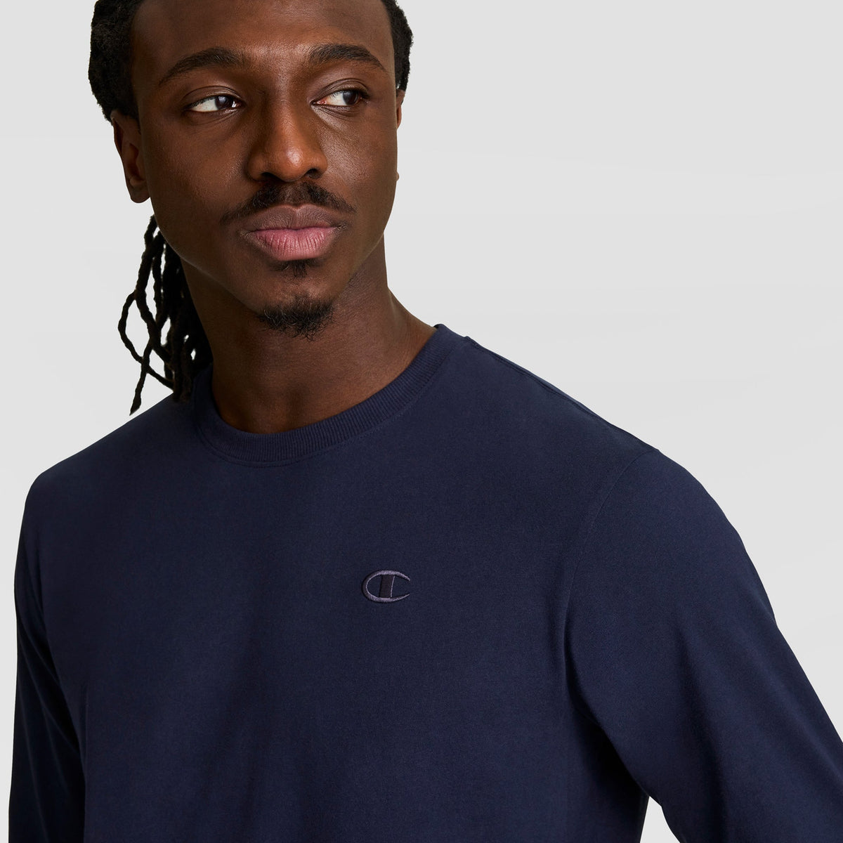 A man with long braided hair wears a navy blue Champion Classic Long Sleeve T-Shirt with an embroidered C logo on the chest, looking to his left against a plain light background.