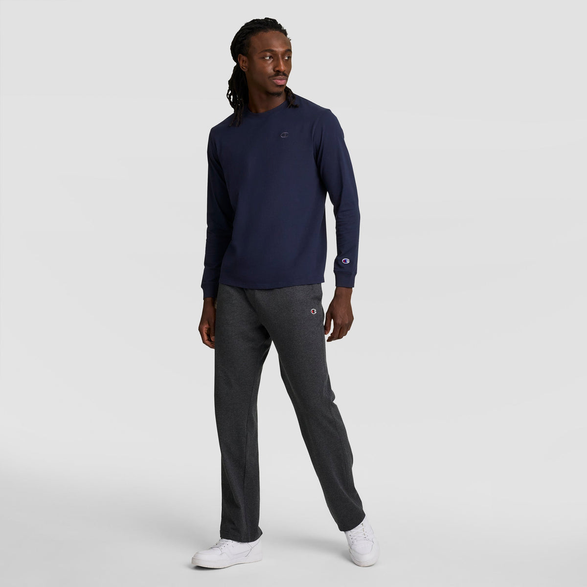 A man stands against a plain background, wearing a navy Champion Classic Long Sleeve T-Shirt with Embroidered C Logo, dark gray sweatpants, and white sneakers. He looks left with one hand by his side and the other on his leg.