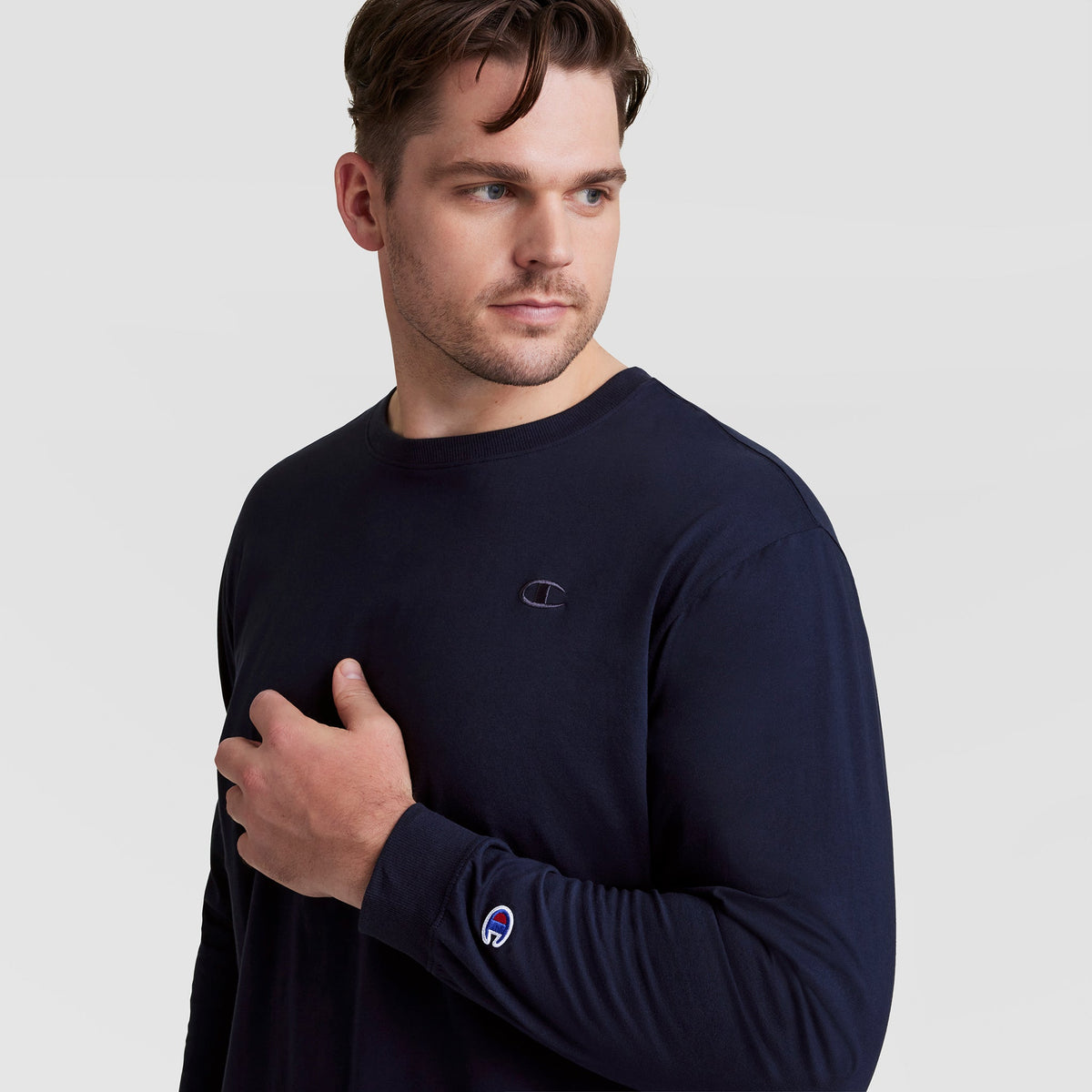 A man with short brown hair wears a Champion Classic Long Sleeve T-Shirt, Embroidered C Logo - Big & Tall in dark navy. He gazes to the side, left hand on his chest, against a plain light background.