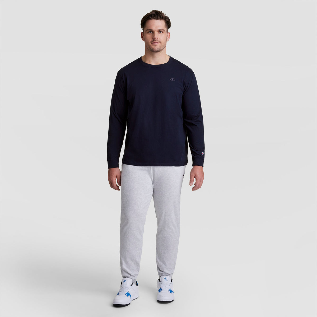 A man stands facing forward wearing a navy Champion Classic Long Sleeve T-Shirt with an embroidered C logo (Big & Tall), light gray sweatpants, and white sneakers with blue accents, against a plain light gray background.