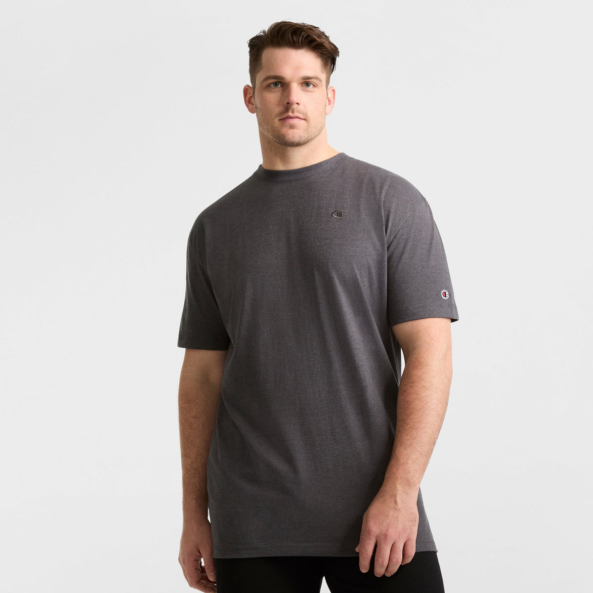 A man in a Champion Classic T-Shirt, C Logo - Big & Tall, dark grey and loose-fitting, with black pants stands against a plain white background, looking at the camera with a neutral expression.
