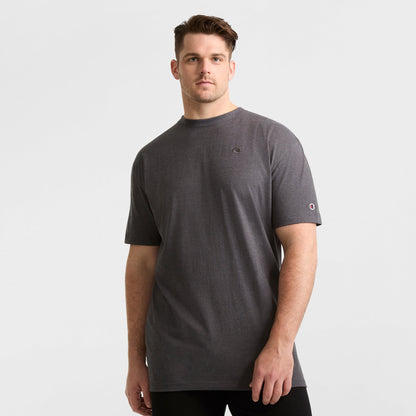A man in a Champion Classic T-Shirt, C Logo - Big & Tall, dark grey and loose-fitting, with black pants stands against a plain white background, looking at the camera with a neutral expression.