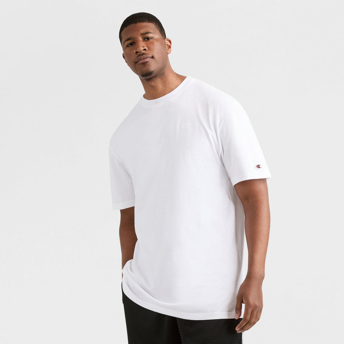 A man with short hair and a neutral expression stands against a plain light background, wearing the Champion Classic T-Shirt, C Logo - Big & Tall in soft cotton and black shorts. One arm is relaxed at his side; the other is slightly bent.