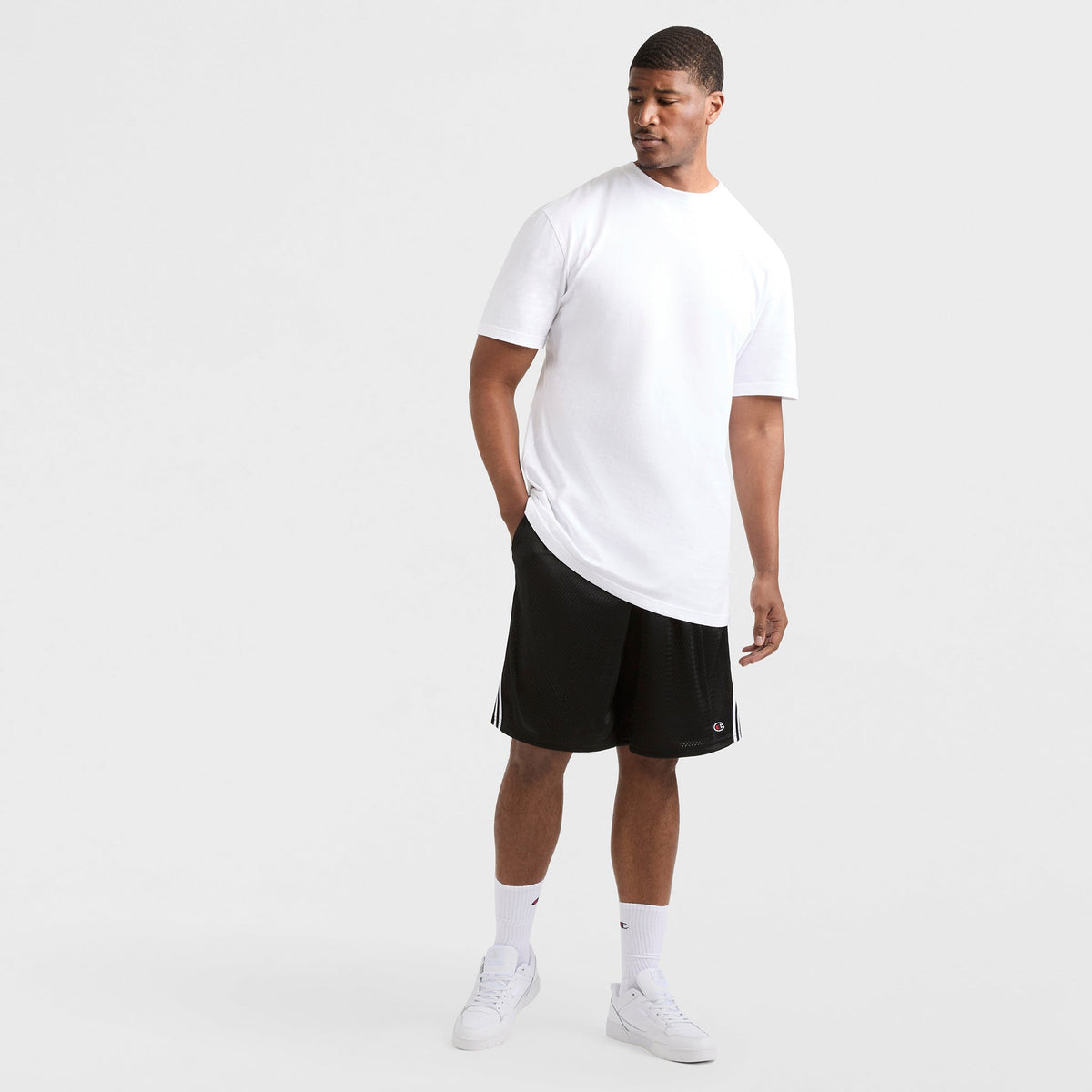 A man stands against a white background wearing a Champion Classic T-Shirt, C Logo - Big & Tall, paired with black athletic shorts with white stripes, white crew socks, and sneakers. One hand is in his pocket as he looks to his left.