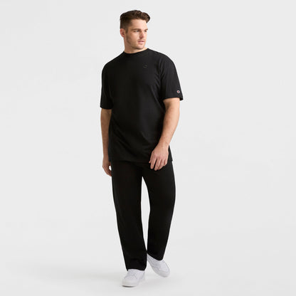 A man wears the Champion Classic T-Shirt, C Logo - Big & Tall in black, styled with matching pants and white sneakers, standing against a white background and gazing to the side with relaxed posture.