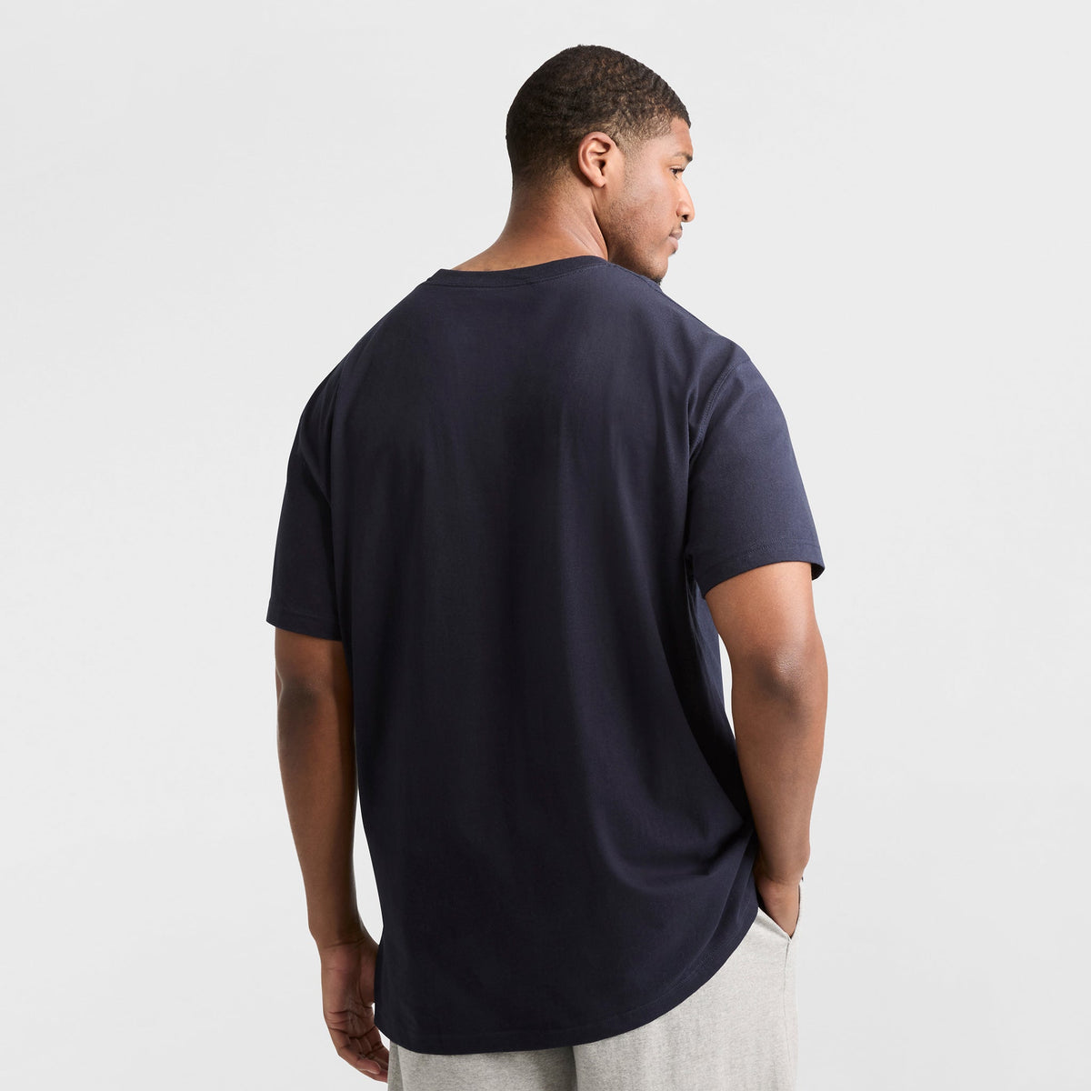 A man stands with his back to the camera, wearing a navy Champion Classic T-Shirt, C Logo - Big & Tall, and light gray pants against a plain white background.