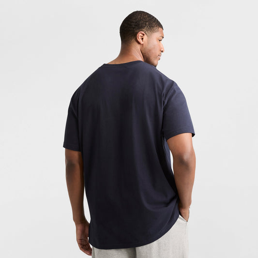 A man stands with his back to the camera, wearing a navy Champion Classic T-Shirt, C Logo - Big & Tall, and light gray pants against a plain white background.