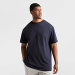 A man wears a Champion Classic T-Shirt, C Logo - Big & Tall in navy blue with light gray pants, standing against a white background and looking aside with one hand in his pocket.