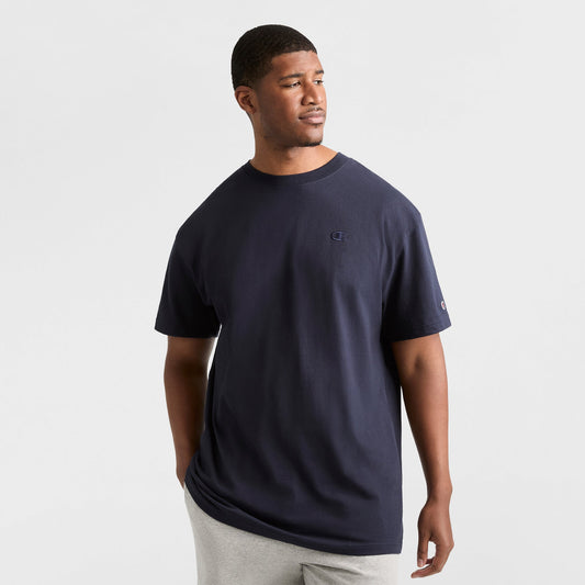 A man wears a Champion Classic T-Shirt, C Logo - Big & Tall in navy blue with light gray pants, standing against a white background and looking aside with one hand in his pocket.