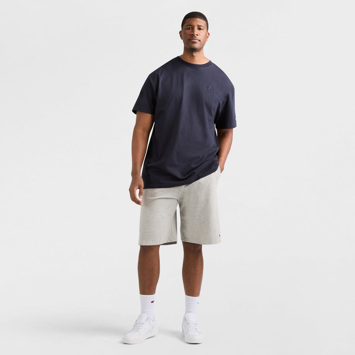 A man stands against a white background wearing the Champion Classic T-Shirt, C Logo - Big & Tall in navy, paired with light gray knee-length shorts, white socks, and white sneakers. One hand is in his pocket and he looks relaxed.