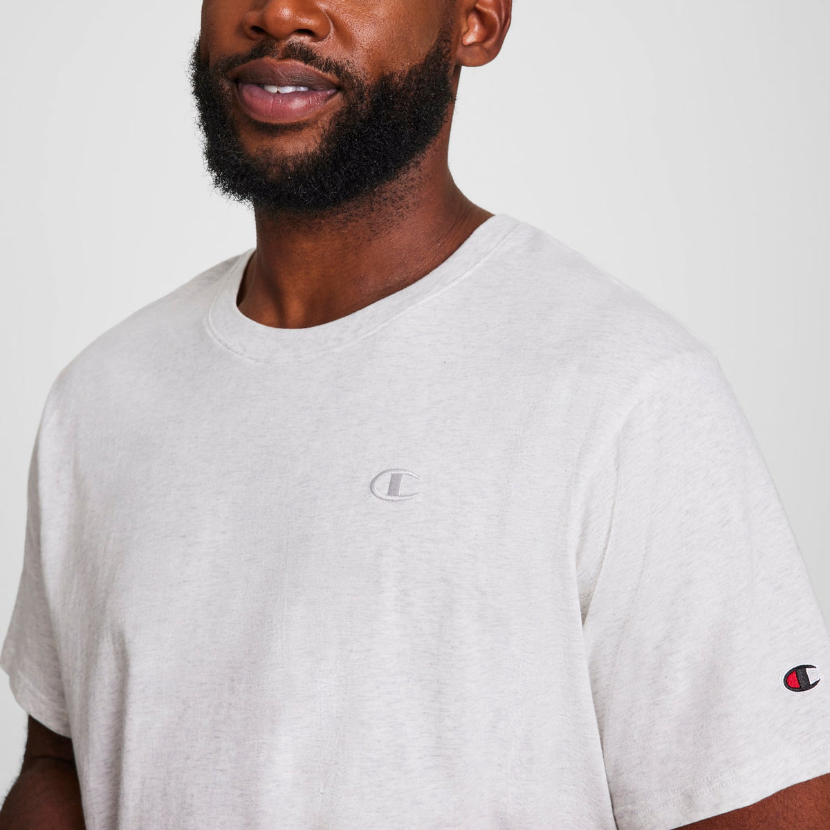 A bearded man wears a light gray Champion Classic T-Shirt, C Logo - Big & Tall, featuring small Champion logos on the chest and left sleeve, posed against a plain background.
