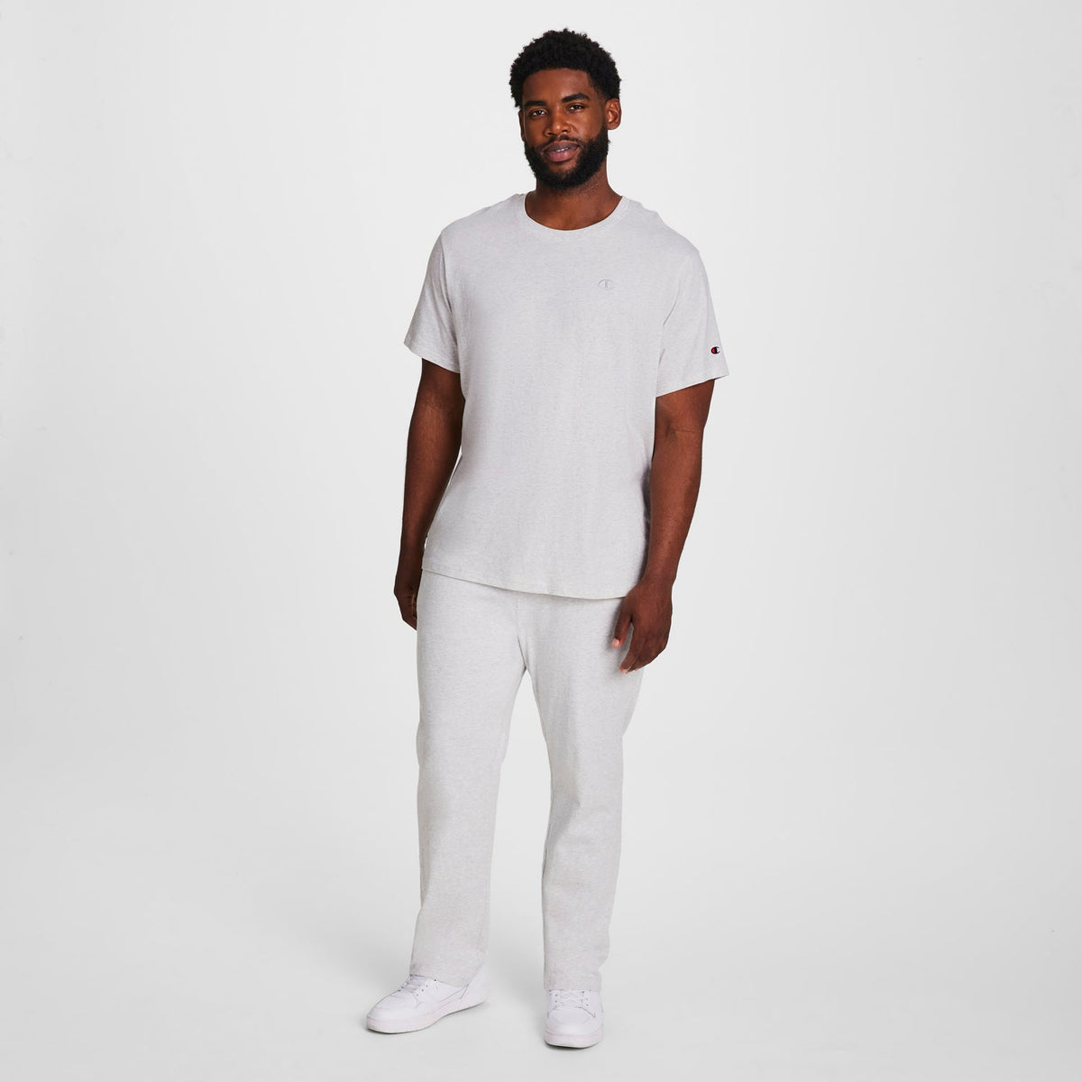 A man with short curly hair and a beard stands against a white background, wearing the Champion Classic T-Shirt, C Logo - Big & Tall in light gray, paired with matching sweatpants and white sneakers. His arms are relaxed by his sides.