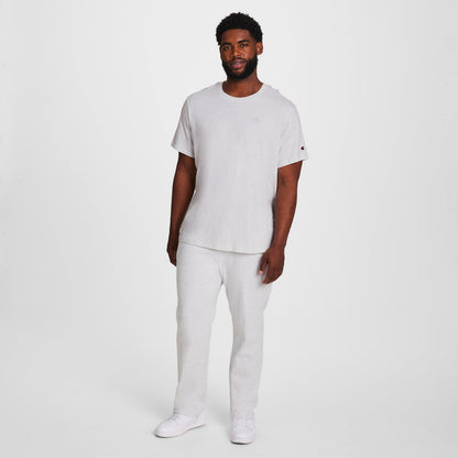 A man with short curly hair and a beard stands against a white background, wearing the Champion Classic T-Shirt, C Logo - Big & Tall in light gray, paired with matching sweatpants and white sneakers. His arms are relaxed by his sides.