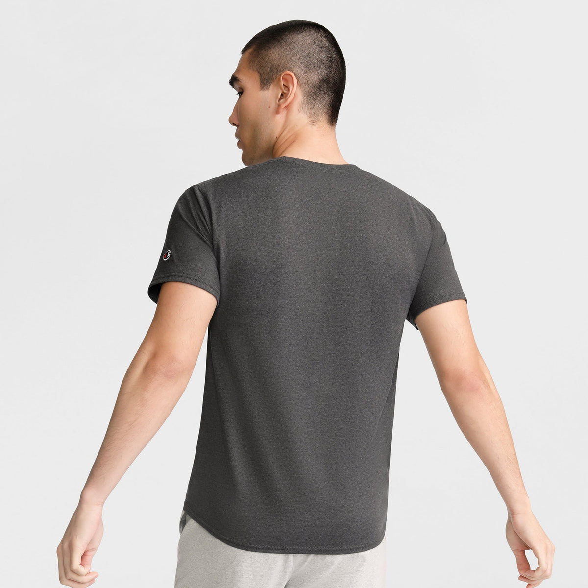 A man with short dark hair stands with his back to the camera, wearing a Champion Classic T-Shirt, C Logo in dark gray and light pants against a plain light background.