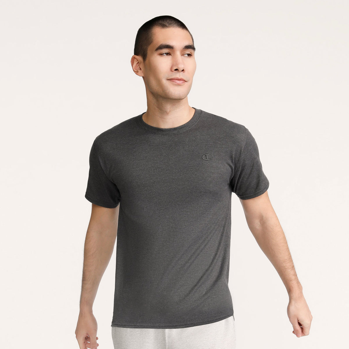 A man with short dark hair wears a Champion Classic T-Shirt, C Logo in dark gray and light gray pants, standing against a light background and looking slightly to his left with a neutral expression.