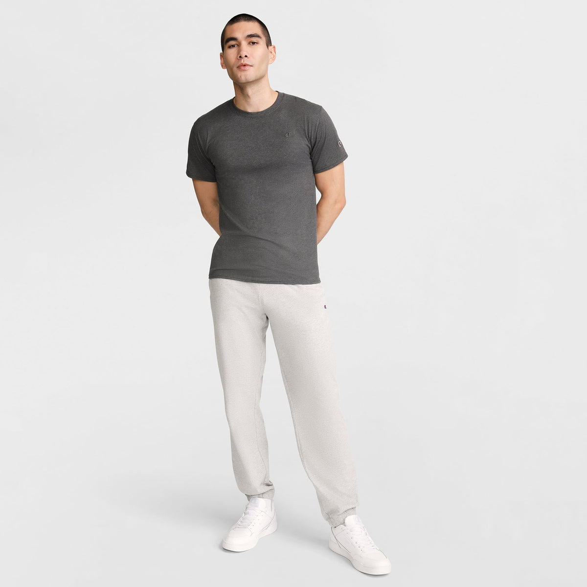 A person stands against a plain white background, wearing a fitted dark gray Champion Classic T-Shirt with C Logo, light gray sweatpants, and white sneakers, hands behind their back.