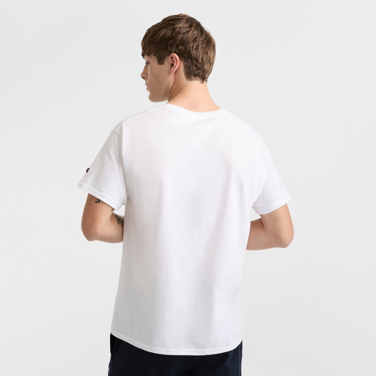 A young man with short brown hair stands with his back to the camera, wearing a Champion Classic T-Shirt, C Logo, and dark pants against a plain white background.