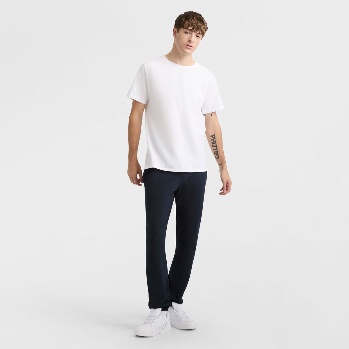 A person stands against a plain white background, wearing the Champion Classic T-Shirt with C Logo, black pants, and white sneakers. With short, tousled hair and a tattoo on their left forearm, they embody classic style.