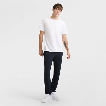 A person stands against a plain white background, wearing the Champion Classic T-Shirt with C Logo, black pants, and white sneakers. With short, tousled hair and a tattoo on their left forearm, they embody classic style.
