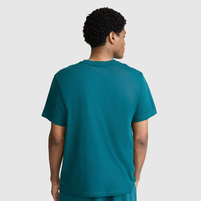 A man with short curly hair is standing with his back to the camera, wearing a Champion Classic T-Shirt, C Logo, paired with matching teal shorts against a plain light gray background.
