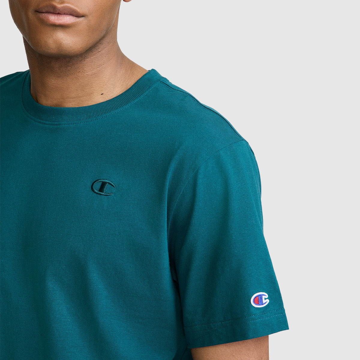 A person wearing a teal Champion Classic T-Shirt, C Logo, featuring the signature Champion branding on the chest and sleeve, stands against a light gray background.