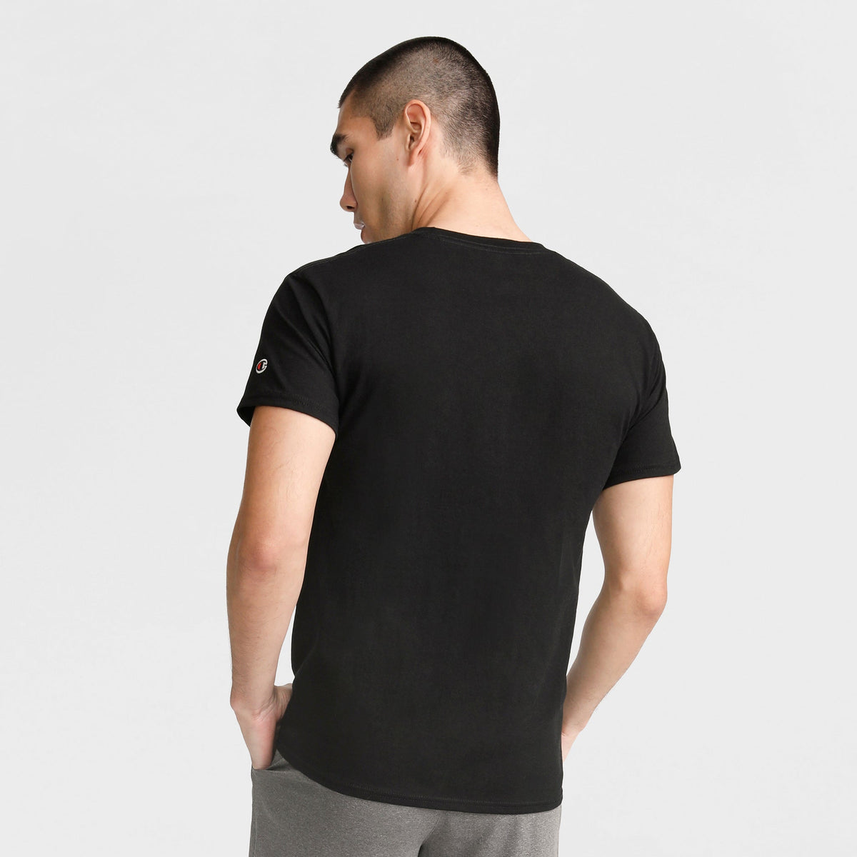 Seen from behind, a person with short hair wears a Champion Classic T-Shirt with C Logo in black cotton and gray pants, standing against a light background with hands in pockets.