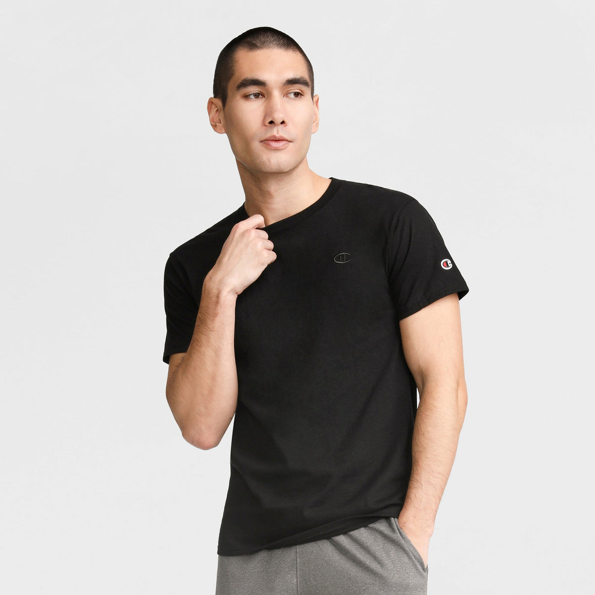 A man wears the Champion Classic T-Shirt, C Logo in plain black with gray pants, standing against a light gray background, looking slightly to the side, one hand on his collar and the other in his pocket.