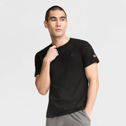A man wears the Champion Classic T-Shirt, C Logo in plain black with gray pants, standing against a light gray background, looking slightly to the side, one hand on his collar and the other in his pocket.