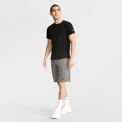 A man stands facing forward in a Champion Classic T-Shirt with C Logo in black, gray shorts, white crew socks, and white sneakers against a plain light background.