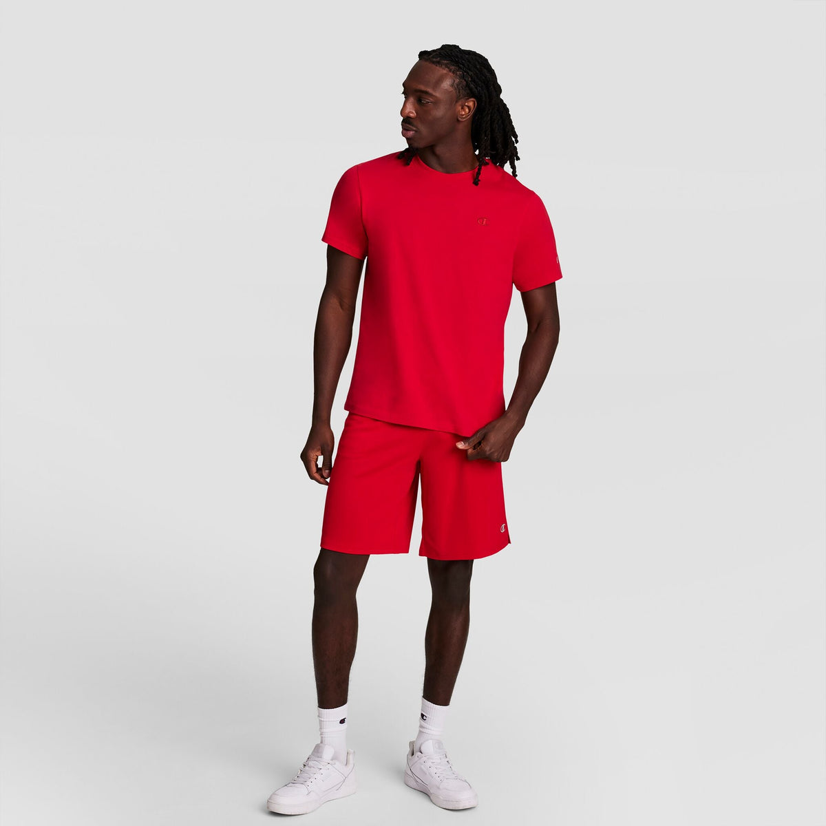 A man with long hair stands against a plain background, wearing a Champion Classic T-Shirt, C Logo in bright red and matching shorts, paired with white socks and sneakers. He looks to his left, one hand by his side, the other touching his shorts.