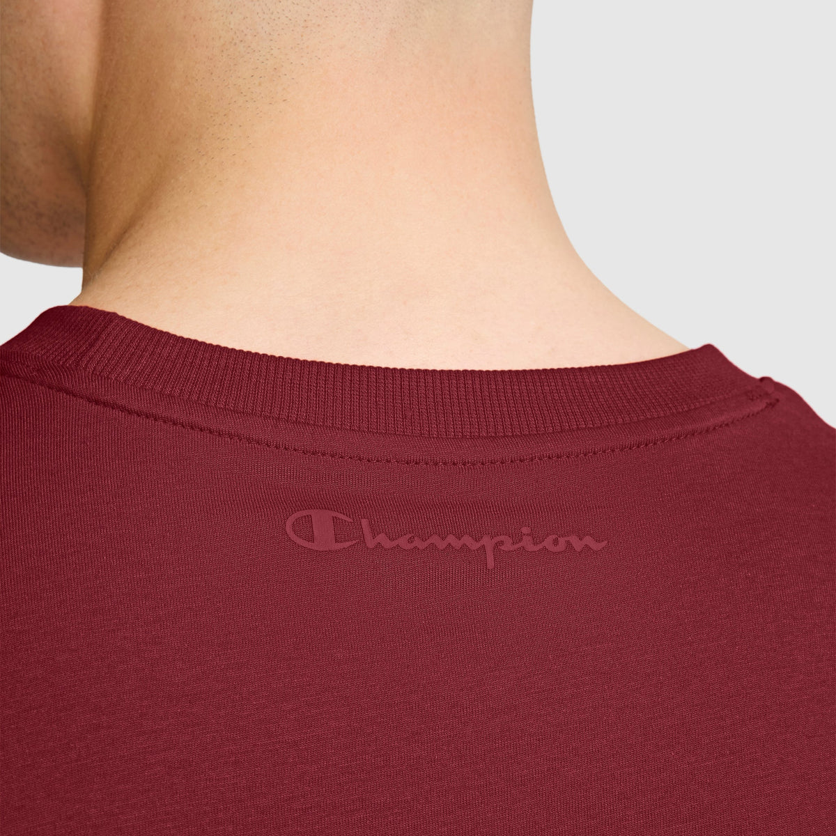 Close-up of the back of a person’s neck and shoulders wearing Champion’s Classic T-Shirt, C Logo in maroon, featuring the tonal Champion logo near the collar.