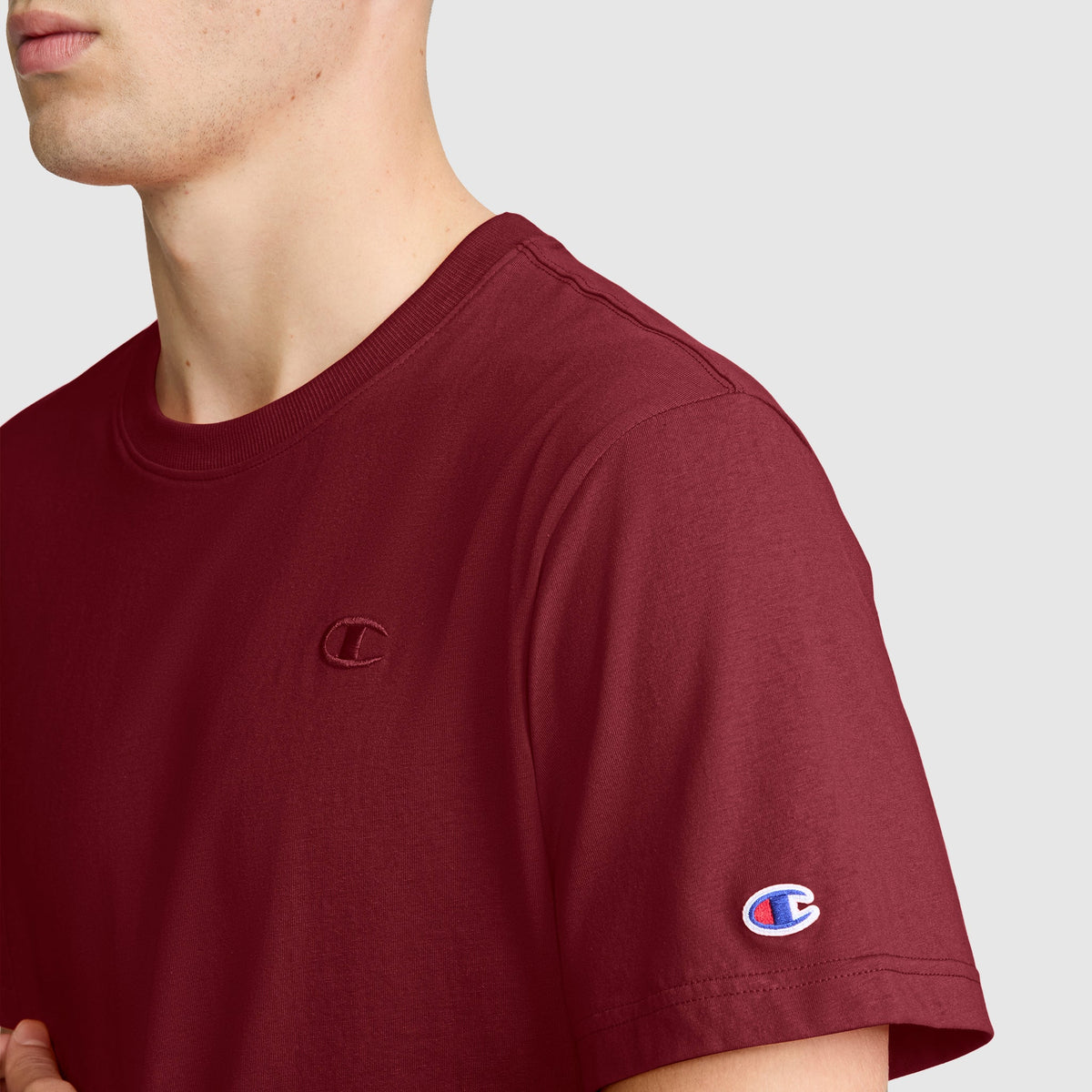 A person is shown from the shoulders up against a plain light background, wearing the Champion Classic T-Shirt, C Logo in maroon. This men’s tee features the brand’s signature embroidered C logo on the chest and sleeve, blending iconic Champion style with everyday comfort.