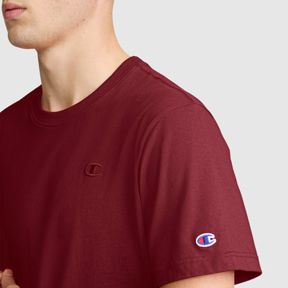 A person is shown from the shoulders up against a plain light background, wearing the Champion Classic T-Shirt, C Logo in maroon. This men’s tee features the brand’s signature embroidered C logo on the chest and sleeve, blending iconic Champion style with everyday comfort.