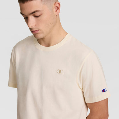 A young man with short hair looks down while wearing Champion's Classic T-Shirt, C Logo, in light beige. The cotton tee displays the iconic Champion logo on both the chest and sleeve against a plain background.