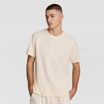 A young man with short hair wears a loose-fitting, light beige Classic T-Shirt with C Logo by Champion and matching pants, standing against a plain light gray background with one hand in his pocket, looking slightly to the side.