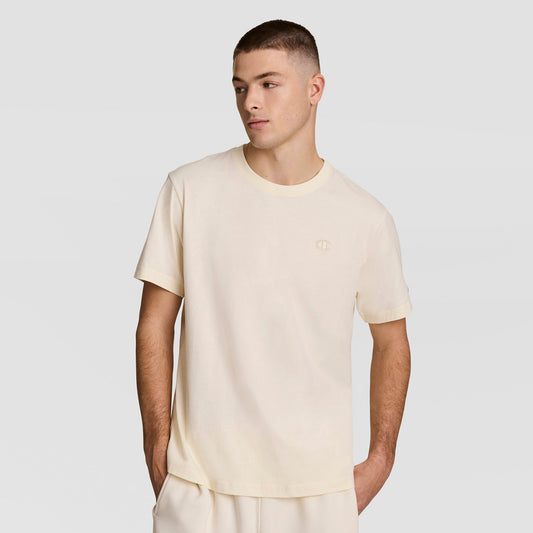 A young man with short hair wears a loose-fitting, light beige Classic T-Shirt with C Logo by Champion and matching pants, standing against a plain light gray background with one hand in his pocket, looking slightly to the side.