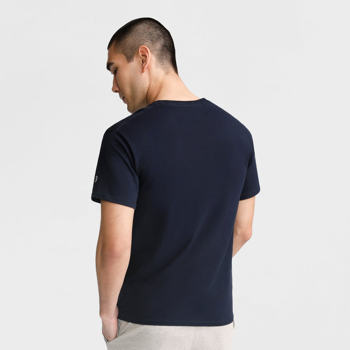 A man with short dark hair, wearing a Champion Classic T-Shirt, C Logo in soft cotton navy blue and light pants, stands with his back to the camera against a plain light background.