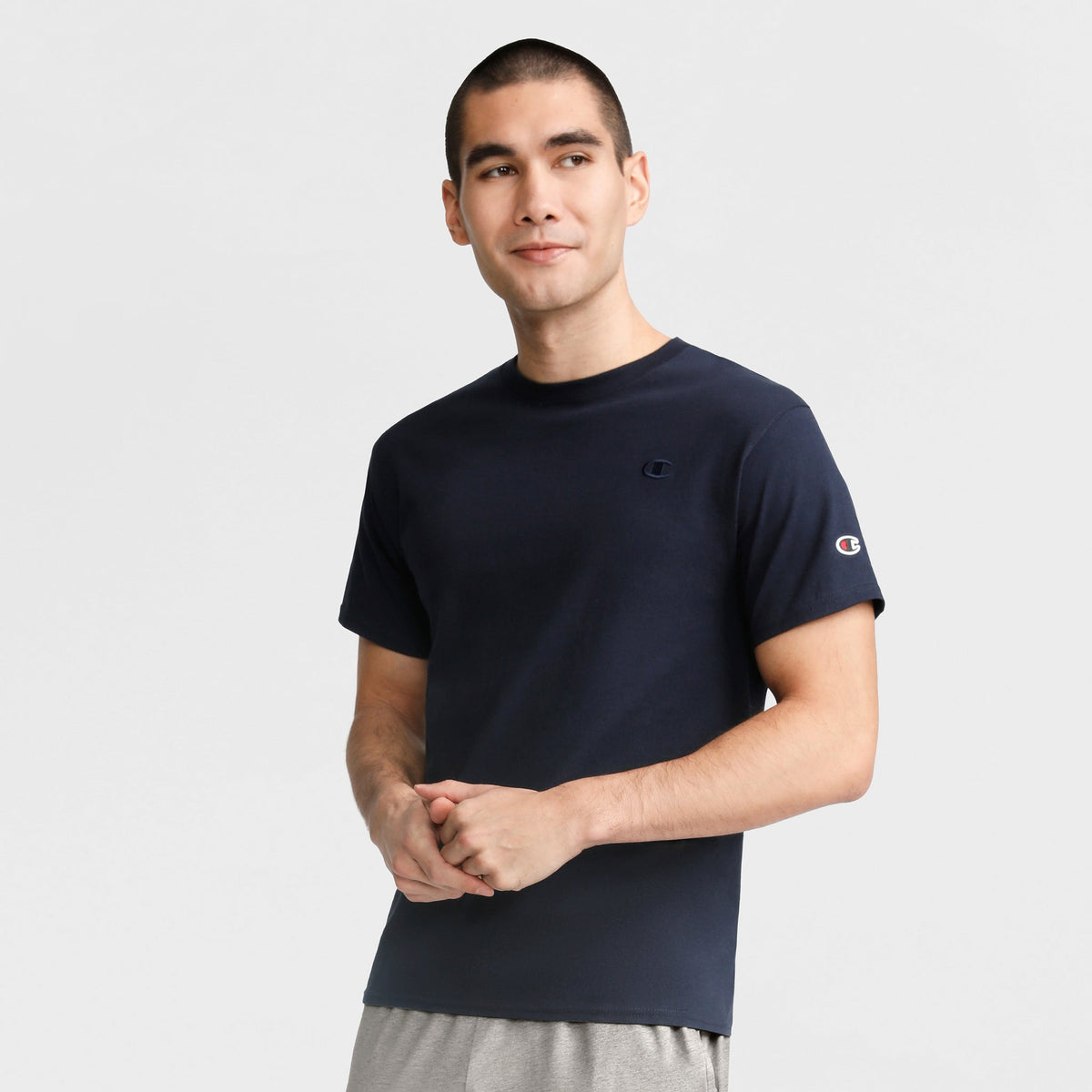 A man with short dark hair wears a Champion Classic T-Shirt, C Logo in navy blue and light gray shorts, standing against a plain white background with a neutral expression, looking slightly to the side.