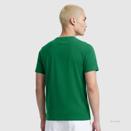 Someone with short, bleached blonde hair is seen from behind wearing a green Champion Classic T-Shirt with the C Logo and white pants, standing against a plain light gray background.
