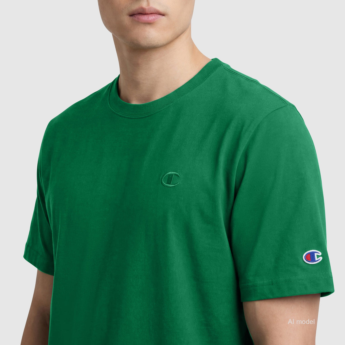 A person wears the Champion Classic T-Shirt, C Logo—a plain green cotton tee with logo on chest and sleeve—against a light grey background. Only their upper body is visible.