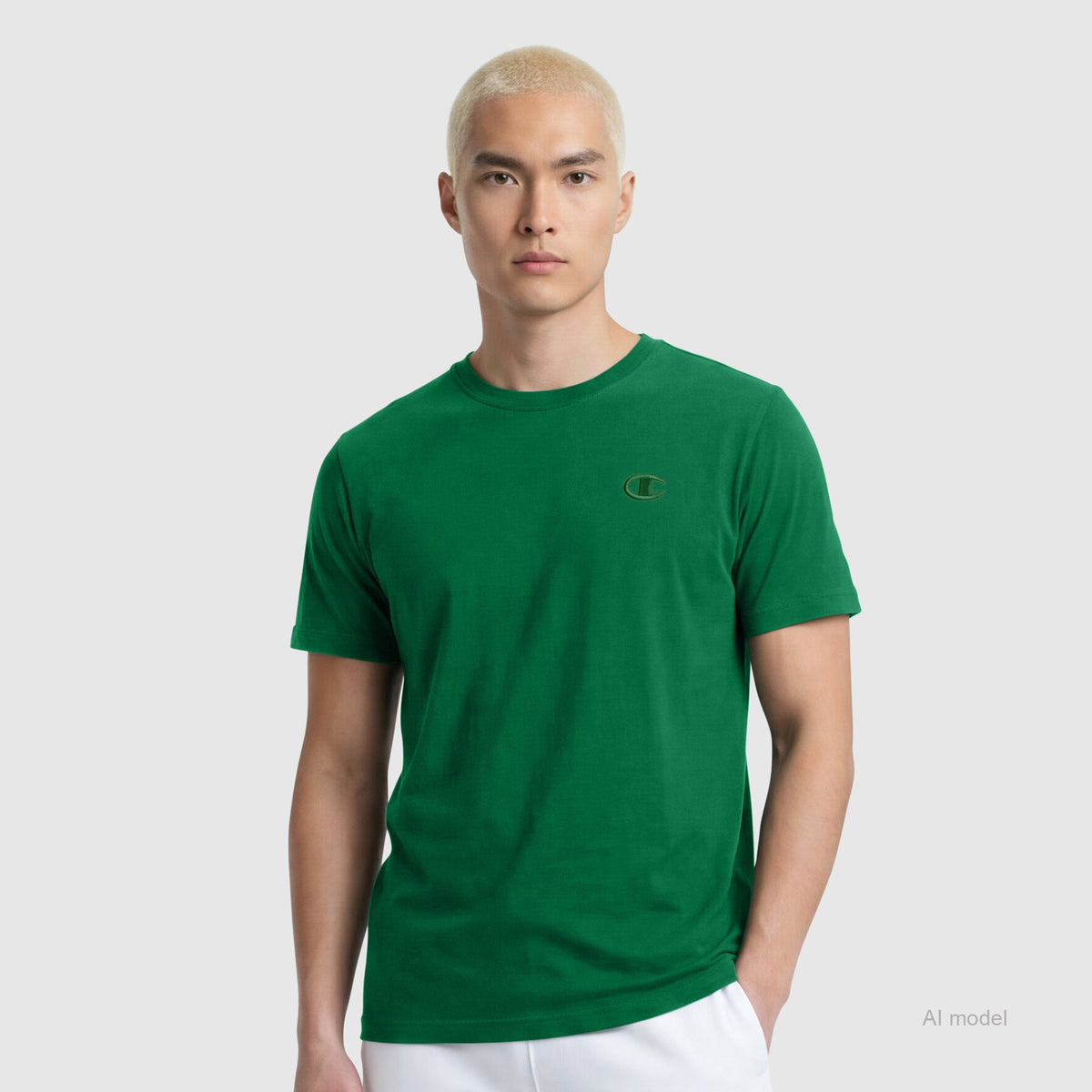 A person with short, bleached blonde hair wears the Champion Classic T-Shirt, C Logo—a plain green cotton tee with a small embroidered logo—paired with white pants, standing against a light gray background.