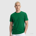 A person with short, bleached blonde hair wears the Champion Classic T-Shirt, C Logo—a plain green cotton tee with a small embroidered logo—paired with white pants, standing against a light gray background.
