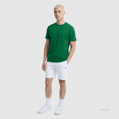 A person with short blond hair wears a green Champion Classic T-Shirt with a C logo, white shorts, white socks, and white sneakers, standing against a plain light gray background.