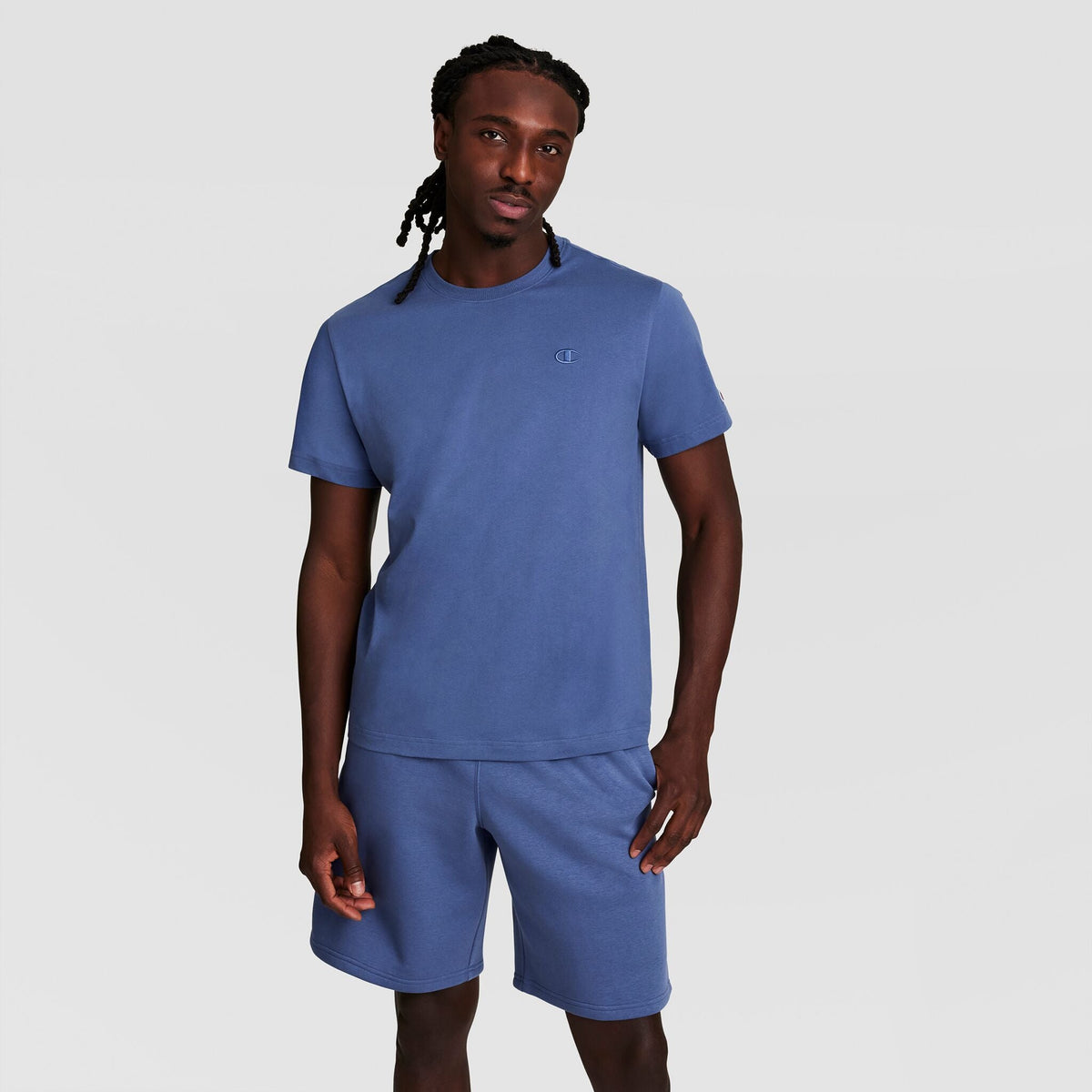 A man with long braided hair wears a matching blue Champion Classic T-Shirt with C Logo and shorts, standing against a plain light gray background.